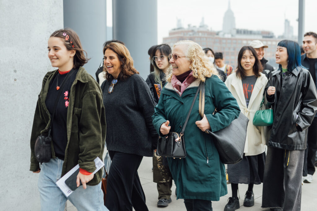A group of people walking outdoors in a city setting. They are dressed in casual, warm clothing. The group appears to be diverse, with a mix of ages and ethnicities. The background includes an urban skyline with tall buildings and cloudy weather. The individuals in the foreground are smiling and engaged in conversation.